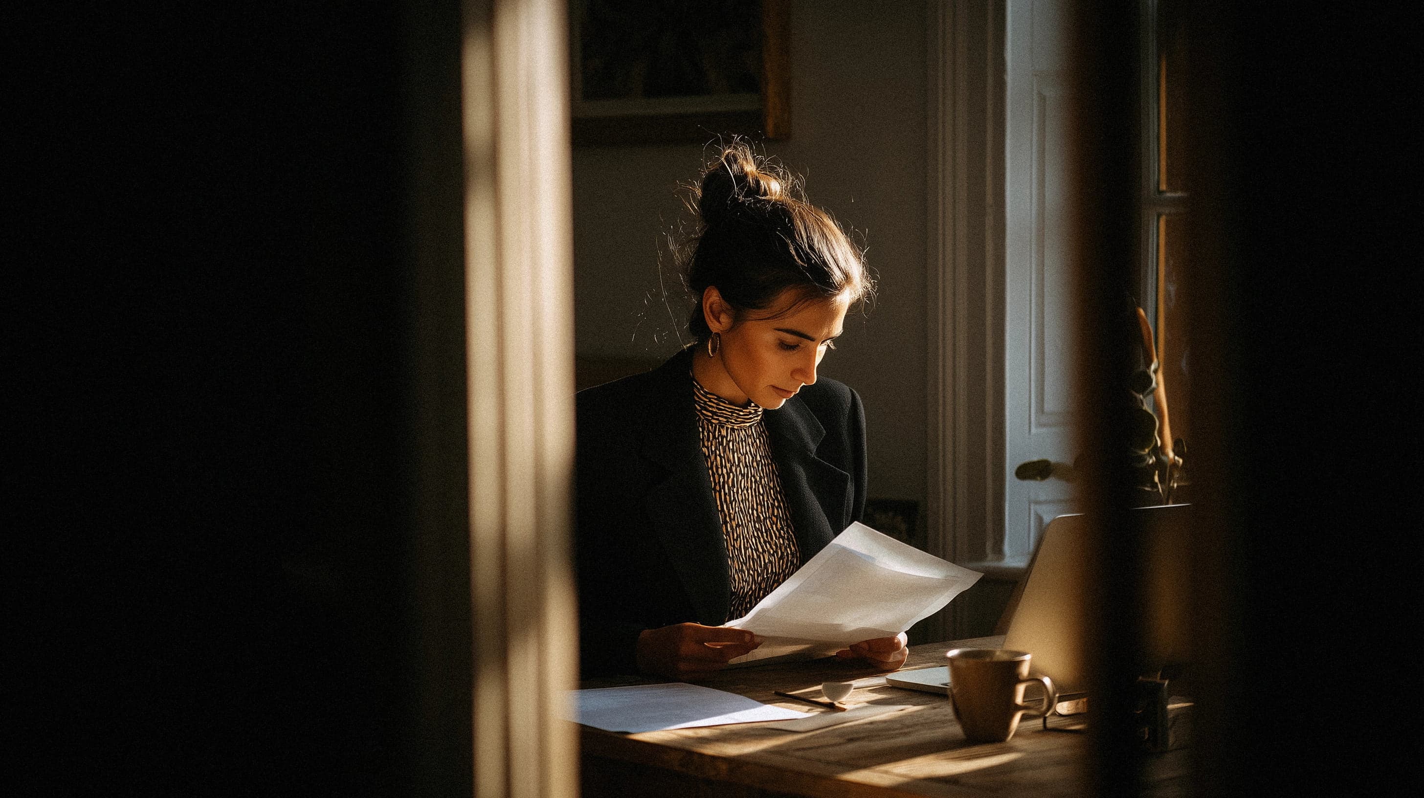 Professional in warm cinematic window light reviewing printed documents at a desk, viewed through a partially open doorway with shallow depth of field